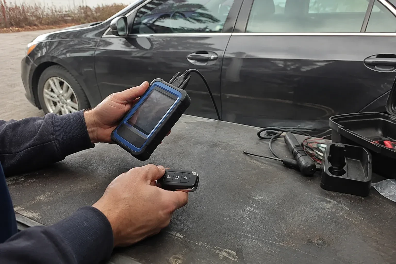 Hands holding a blue-edged electronic device and car-key fob at a worktable beside a parked car. — Transponder Key Programming, Norfolk County
