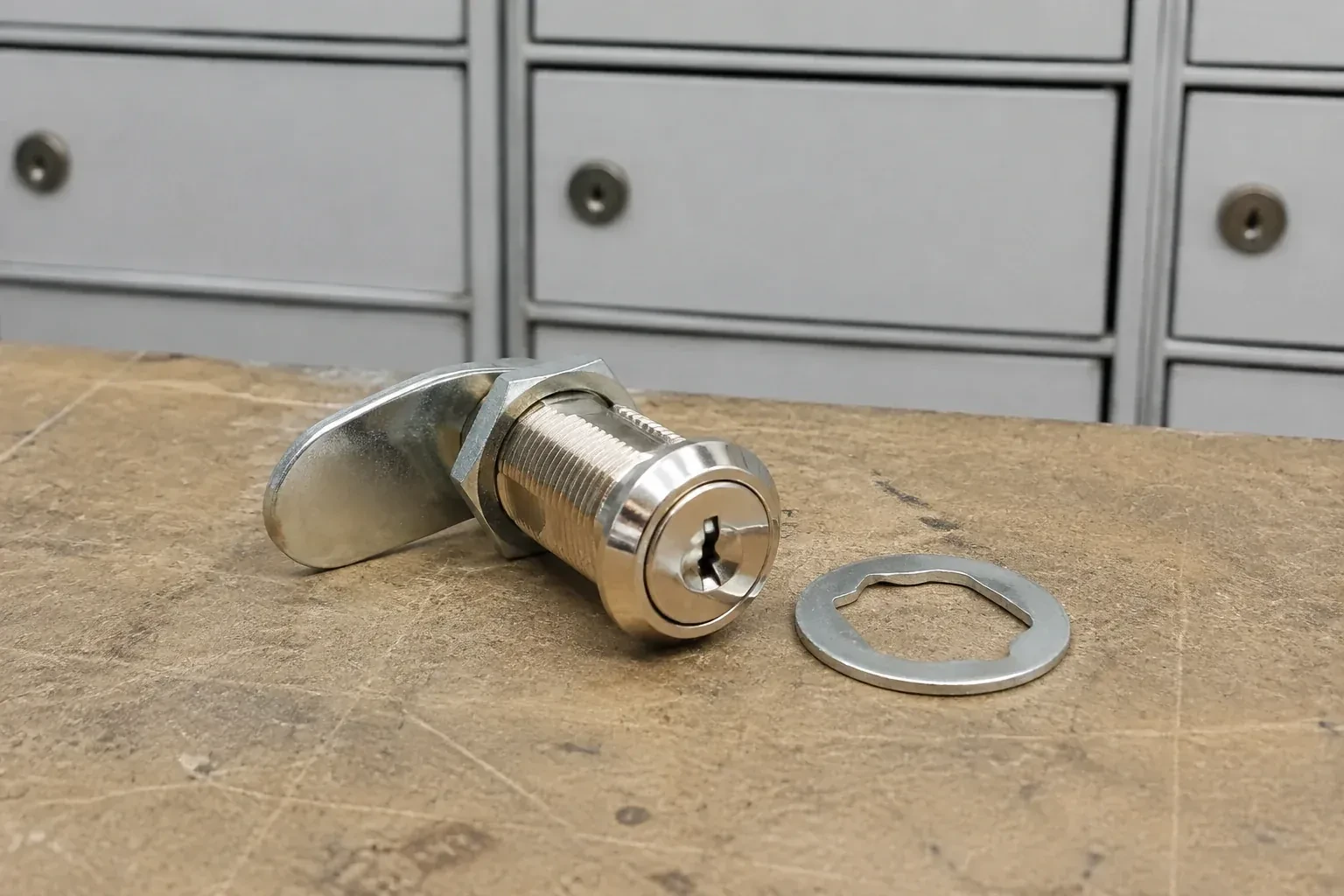 Detached threaded cam lock and mounting washer on a workbench in front of closed mail compartments. — Mailbox Lock Replacement, Norfolk County